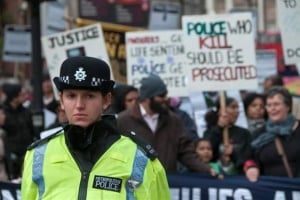 Image shows a police officer in front of a group of people campaigning for greater justice for people killed by police officers.