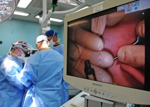 Image shows doctors performing surgery, with a close-up on a monitor in the foreground.