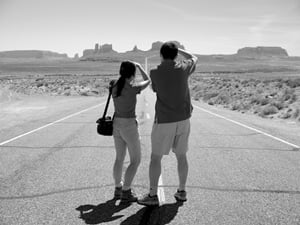 Image shows a black and white photograph of two tourists on a road in the desert, themselves taking photographs. 